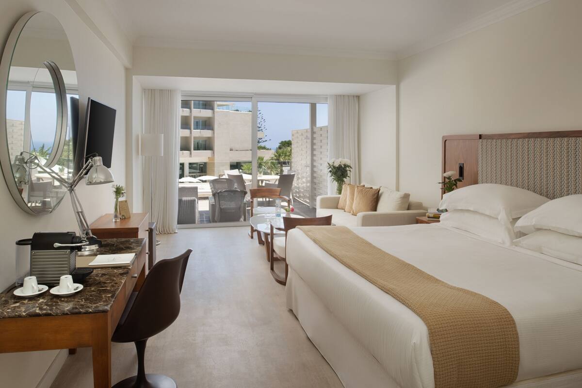 Une chambre d’hôtel avec un grand lit, un bureau avec un ordinateur, une chaise, une petite table avec une plante et une vue sur le balcon extérieur.