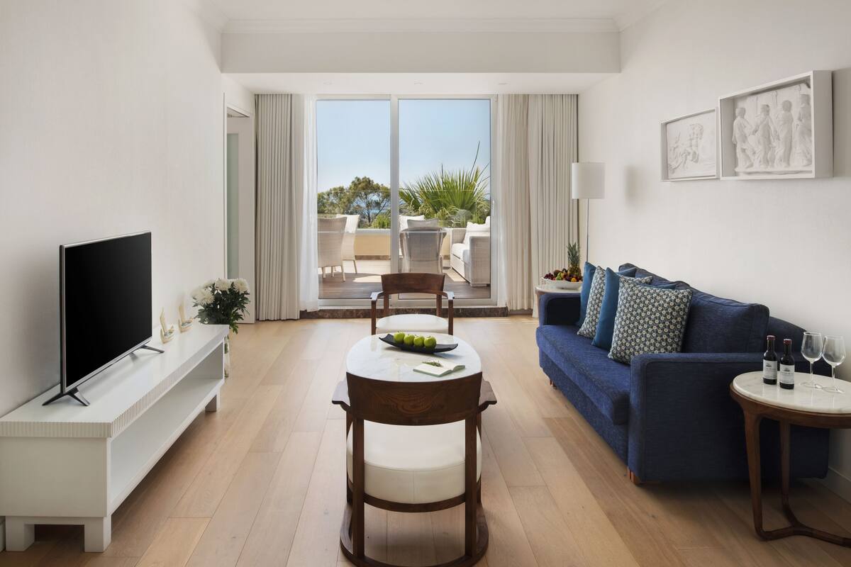 Un salon moderne doté d’un téléviseur à écran plat, d’une table à manger avec des chaises, d’un canapé bleu et d’un balcon aménagé avec du mobilier d’extérieur.