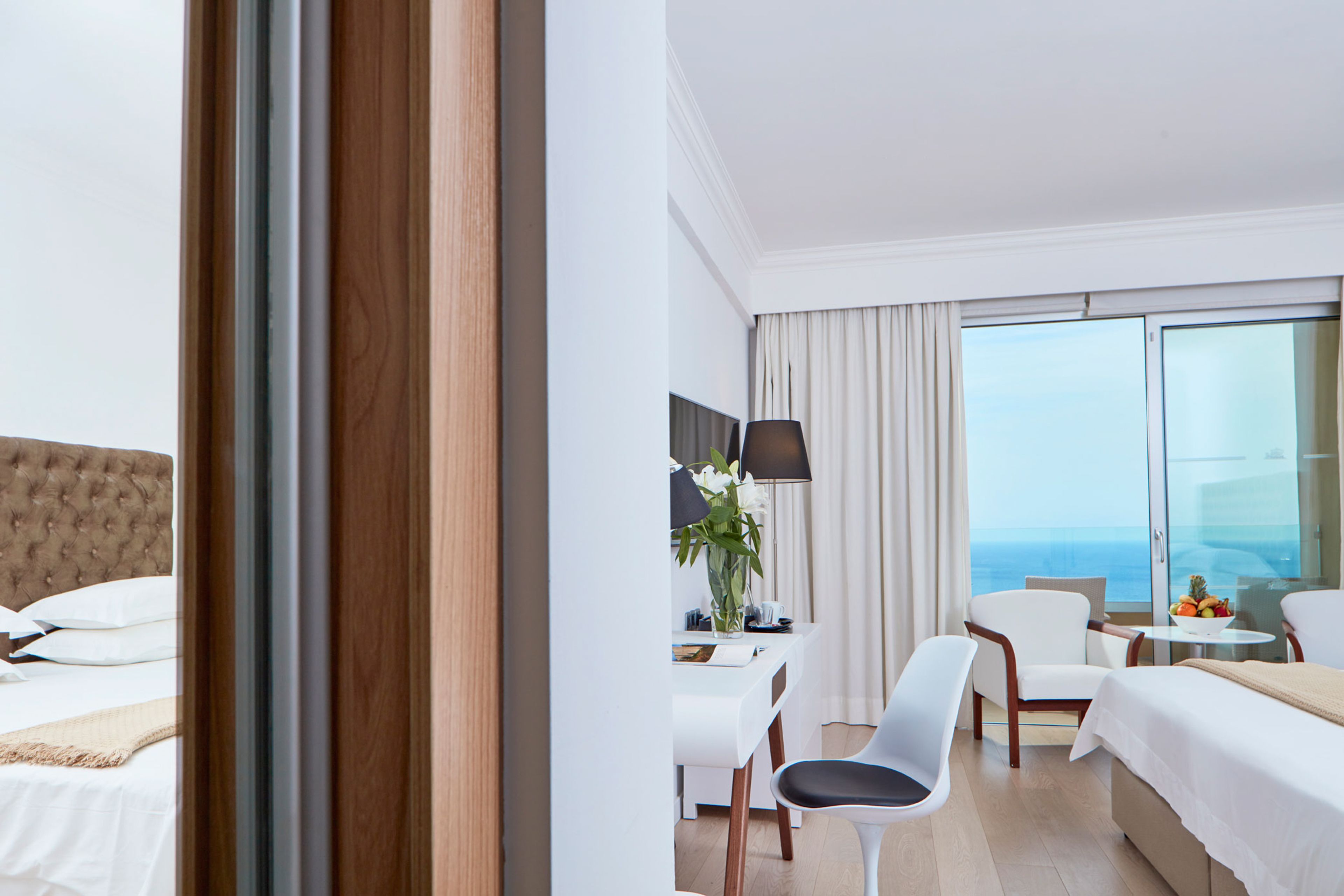 A hotel room with a bed, a desk with a lamp, two chairs, and a view of the ocean.