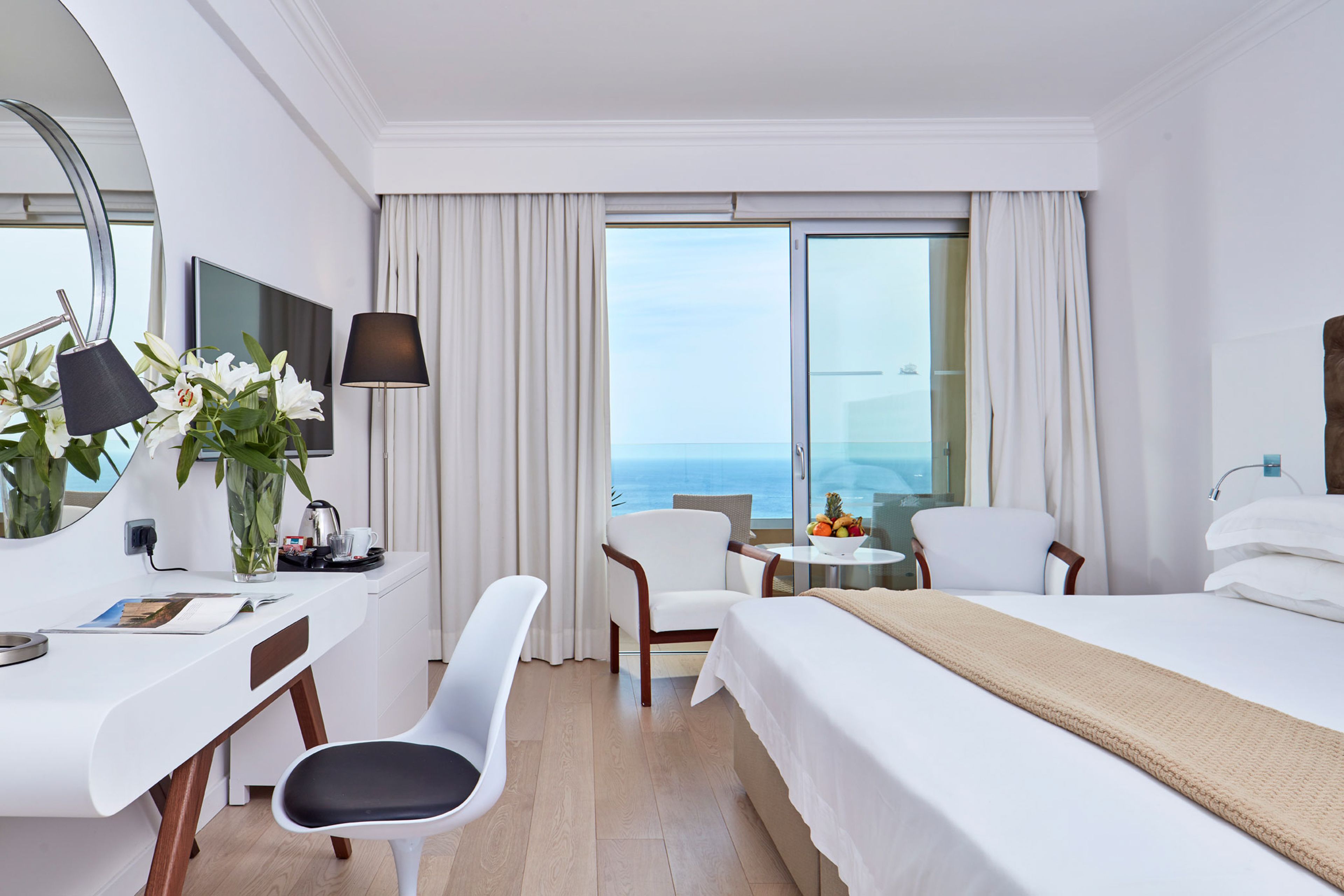 A hotel room with a bed, two chairs, a desk, a TV, and a view of the ocean.