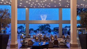A spacious lobby with a grand chandelier, large windows offering a view of the pool and outdoor area, and a seating area with tables and chairs. - Grecian Park Hotel (Paralimni)
