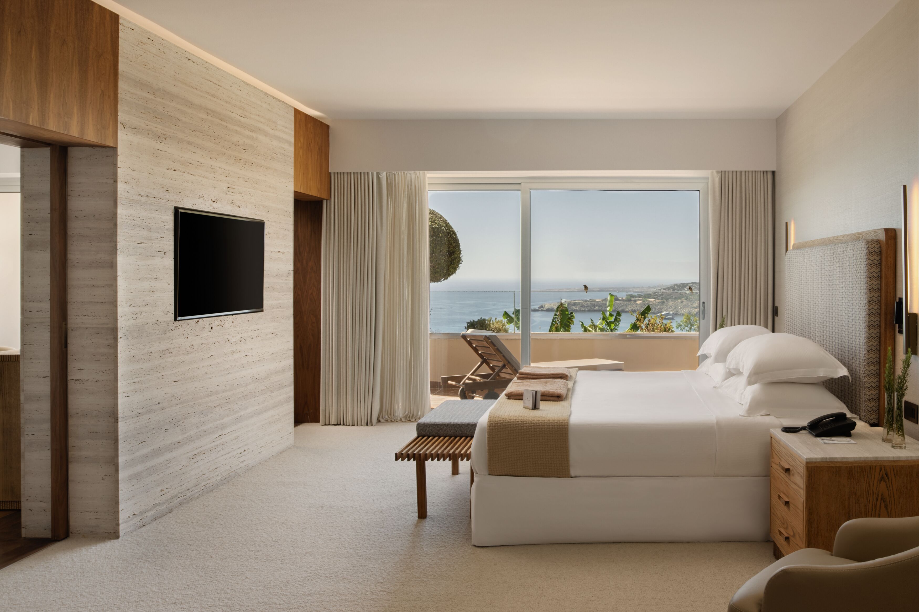 A modern hotel room with a large bed, a TV mounted on the wall, a desk with a chair, and a view of the sea.
