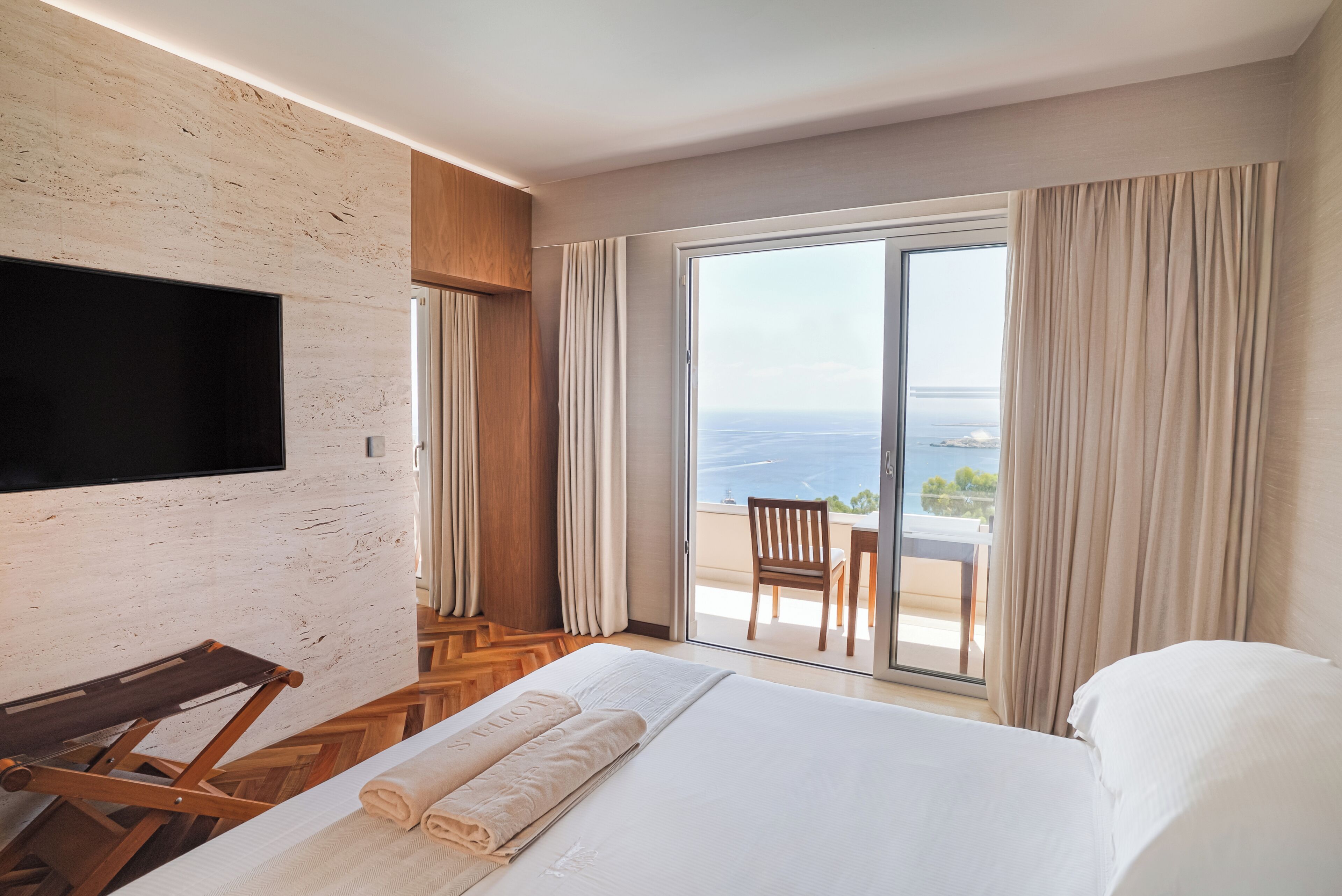 A modern hotel room with a large bed, a flat-screen TV mounted on a textured wall, and a balcony with a view of the sea.