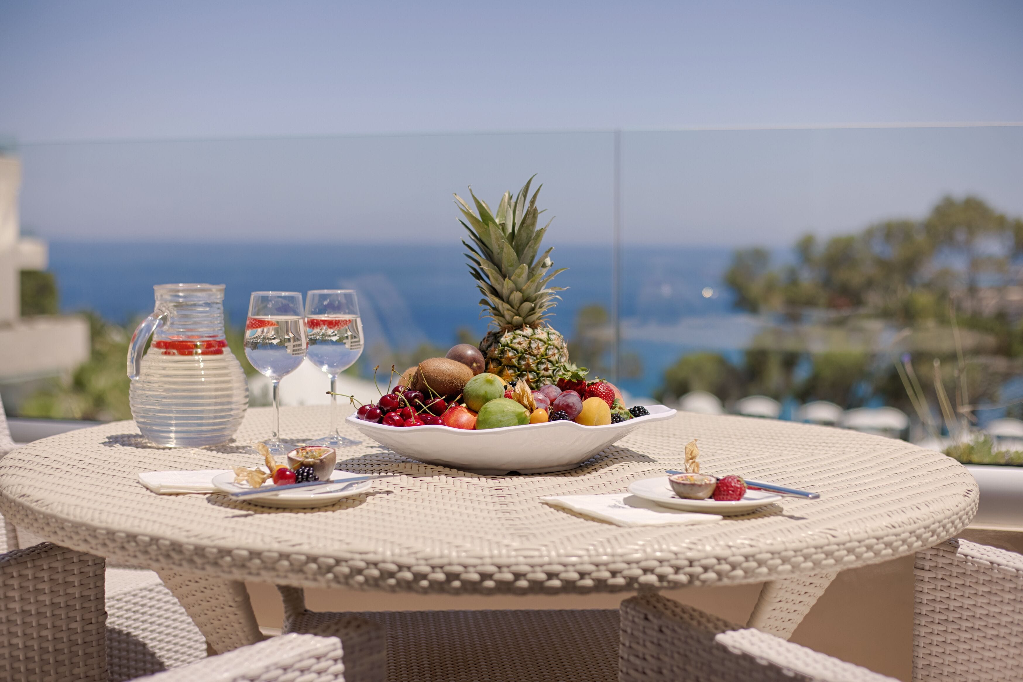Une table ronde en osier, sur laquelle sont disposés un panier de fruits, deux verres, une carafe et une petite assiette avec une cuillère, le tout placé devant un paysage marin pittoresque.