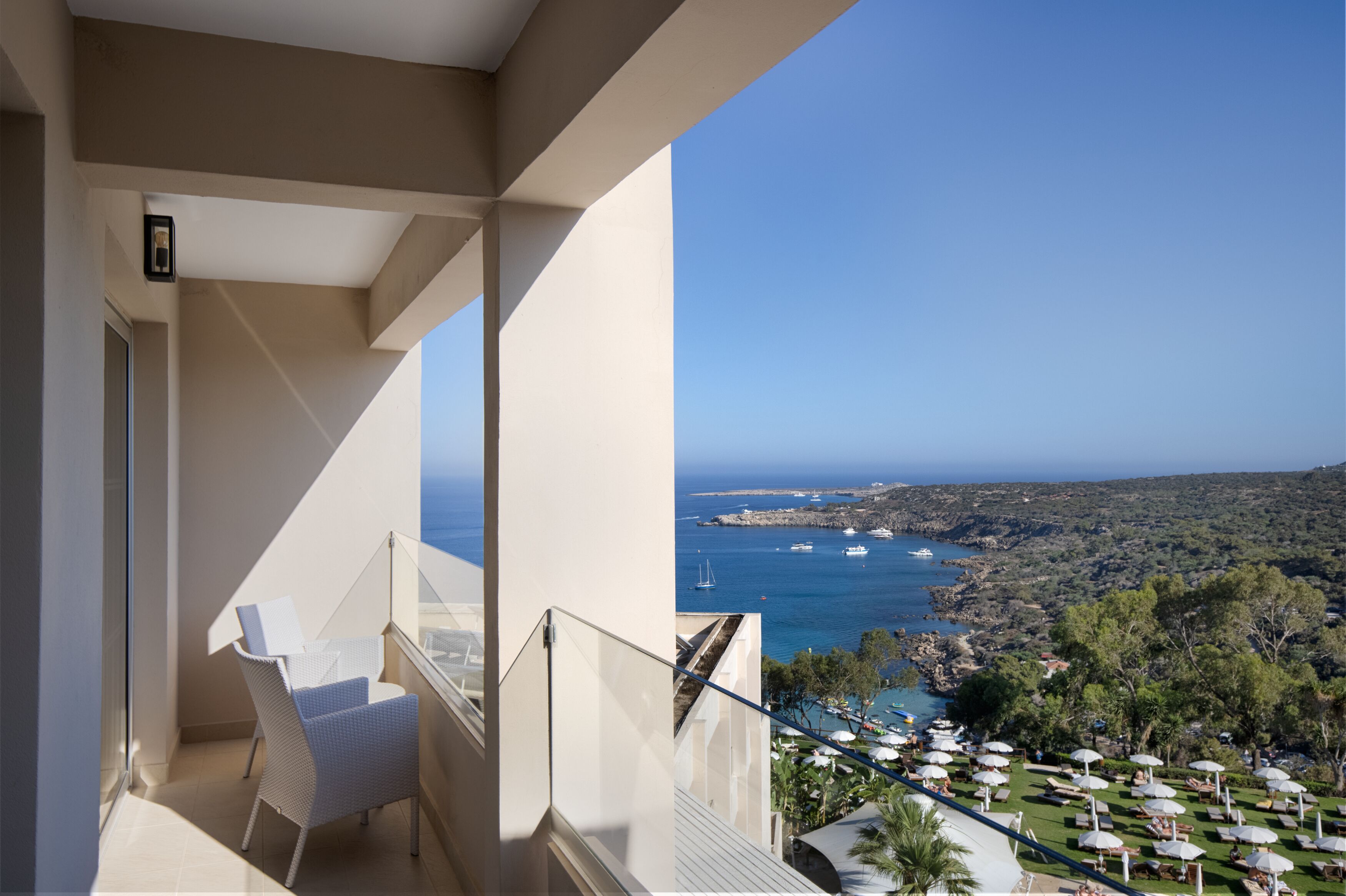 Ein Balkon mit weißem Stuhl, Glasgeländer und Blick auf das Meer und die grüne Landschaft darunter.