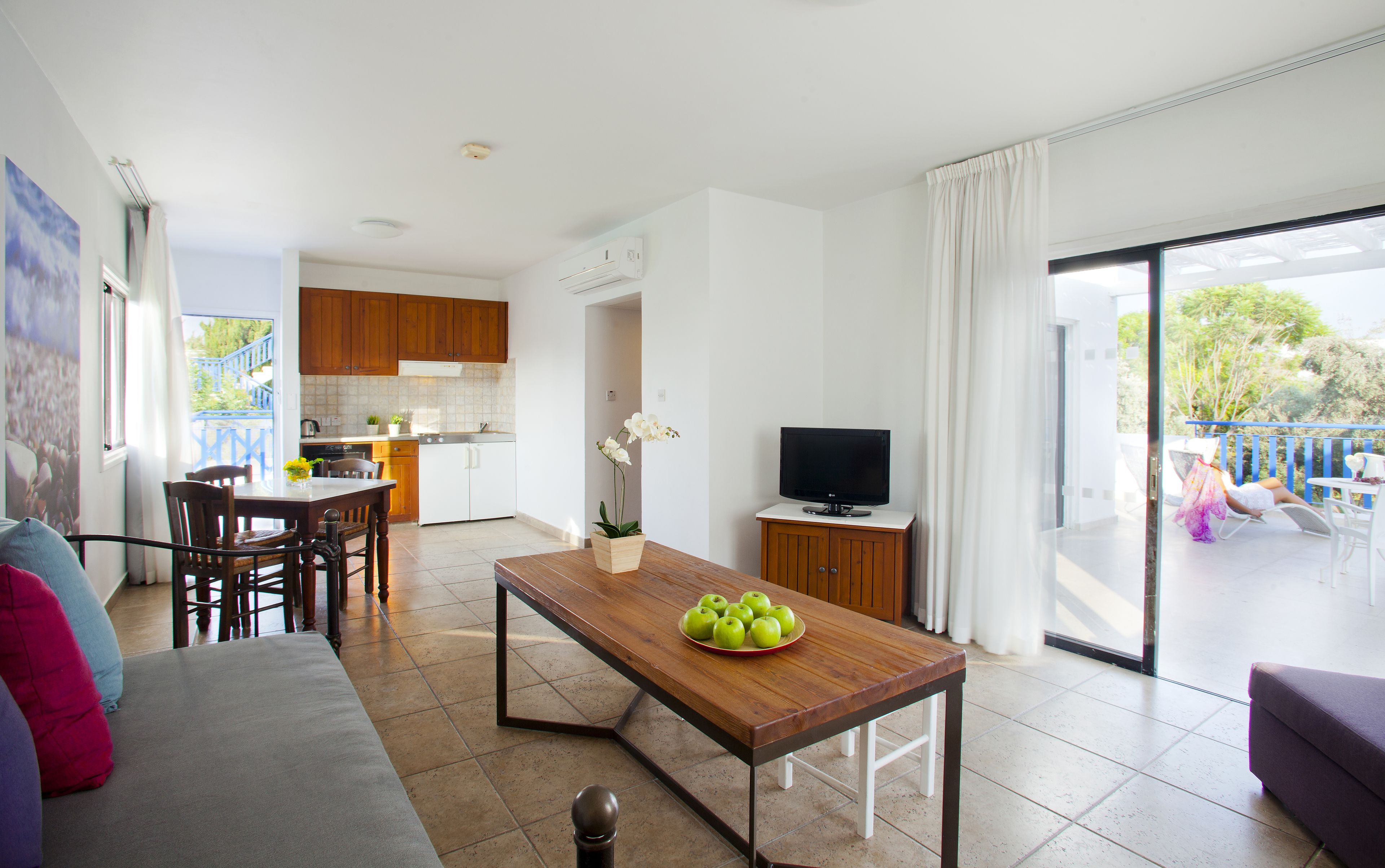 Una sala de estar moderna con una mesa de comedor de madera, un sofá y un televisor. Se observa una cocina al fondo con armarios de madera y un balcón con vistas.