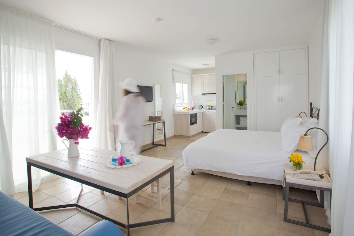 Habitación de hotel con cama, una mesita con un florero, una mesita de noche con lámpara y vistas a la cocina y al balcón.
