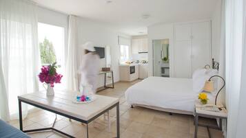 Une chambre d’hôtel avec un lit, une petite table ornée d’un vase de fleurs, une table de chevet avec une lampe, et une vue sur la cuisine et le balcon.