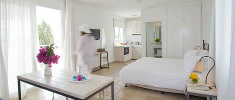 Habitación de hotel con cama, una mesita con un florero, una mesita de noche con lámpara y vistas a la cocina y al balcón.