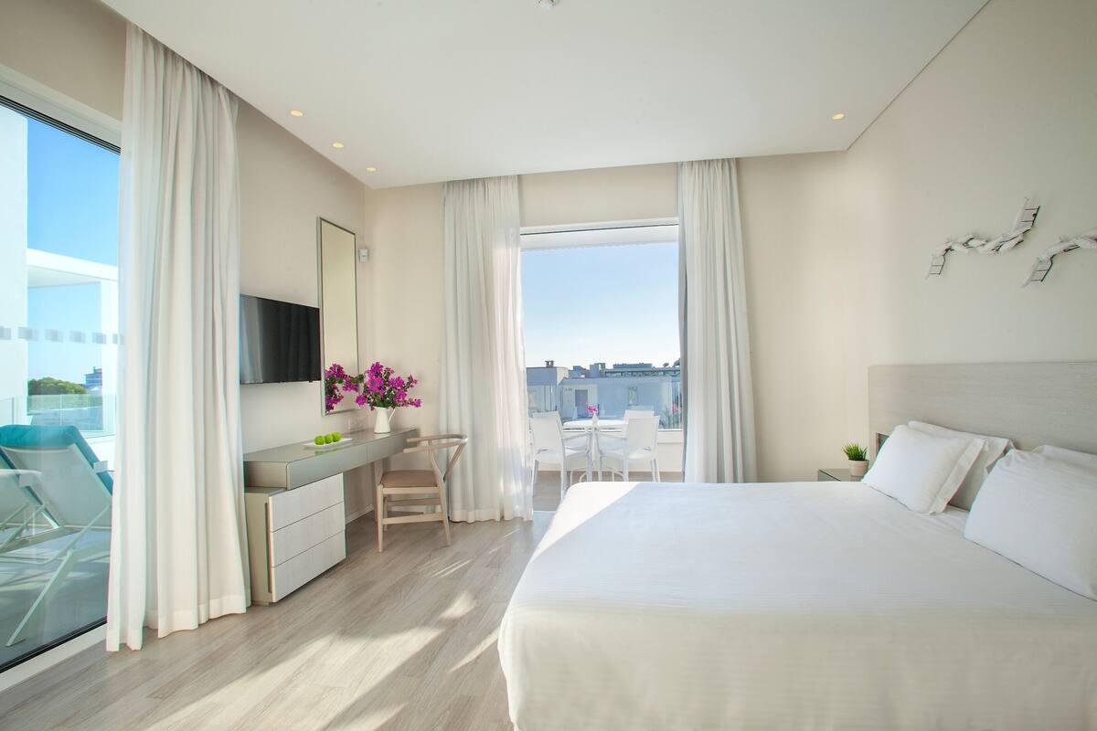 Habitación de hotel moderna con una cama grande, un escritorio, una silla, un televisor y un balcón con vistas a la ciudad.
