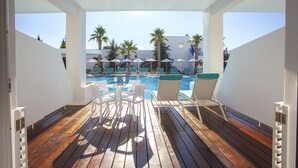 A wooden deck with white chairs and umbrellas overlooking a pool area with palm trees and a building in the background. - Aliathon Aegean (Geroskipou)