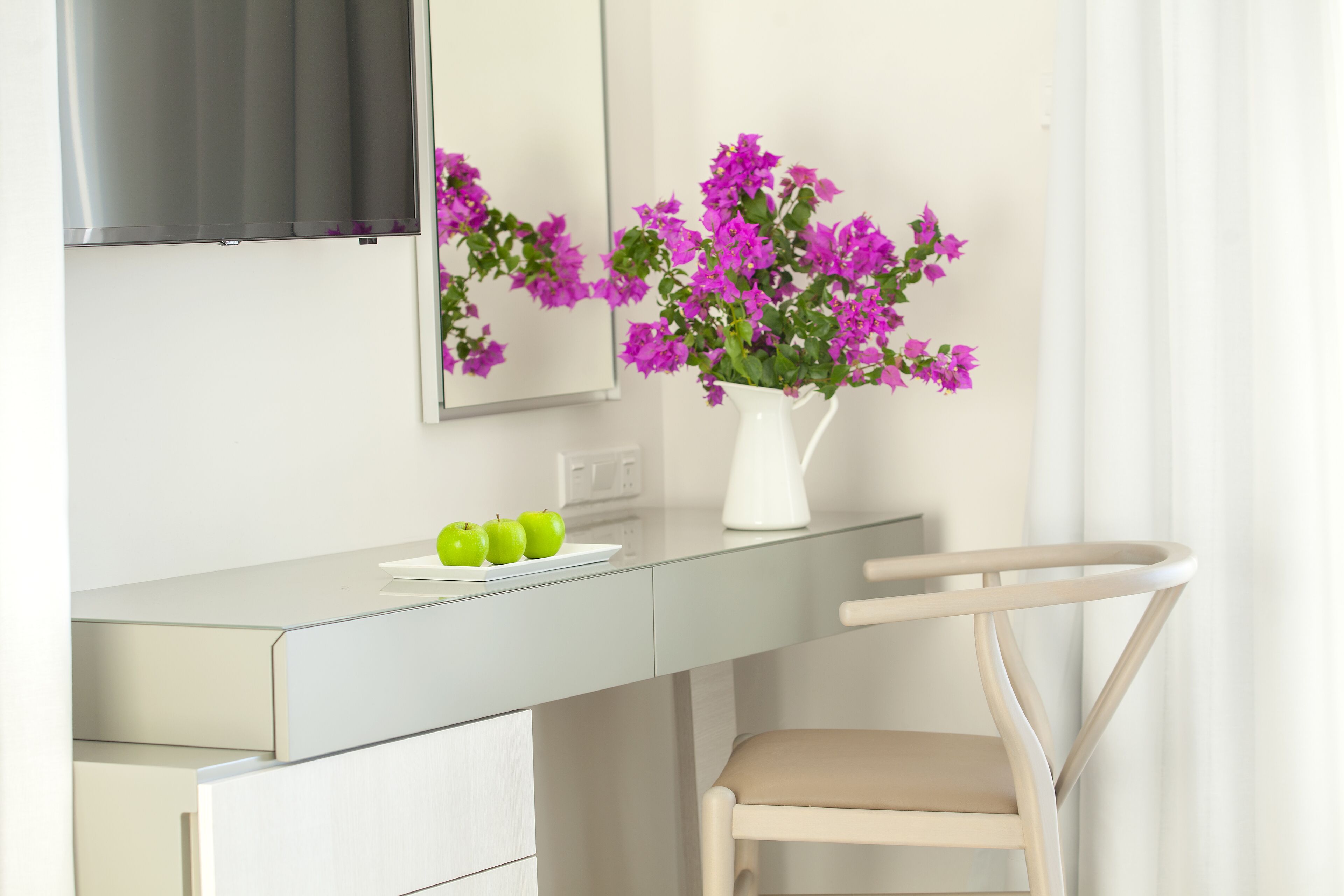 Ein modernes Badezimmer mit einem weißen Waschtisch, einem Spiegel, einem darüber angebrachten Fernseher und einer Vase mit violetten Blumen auf dem Waschtisch.