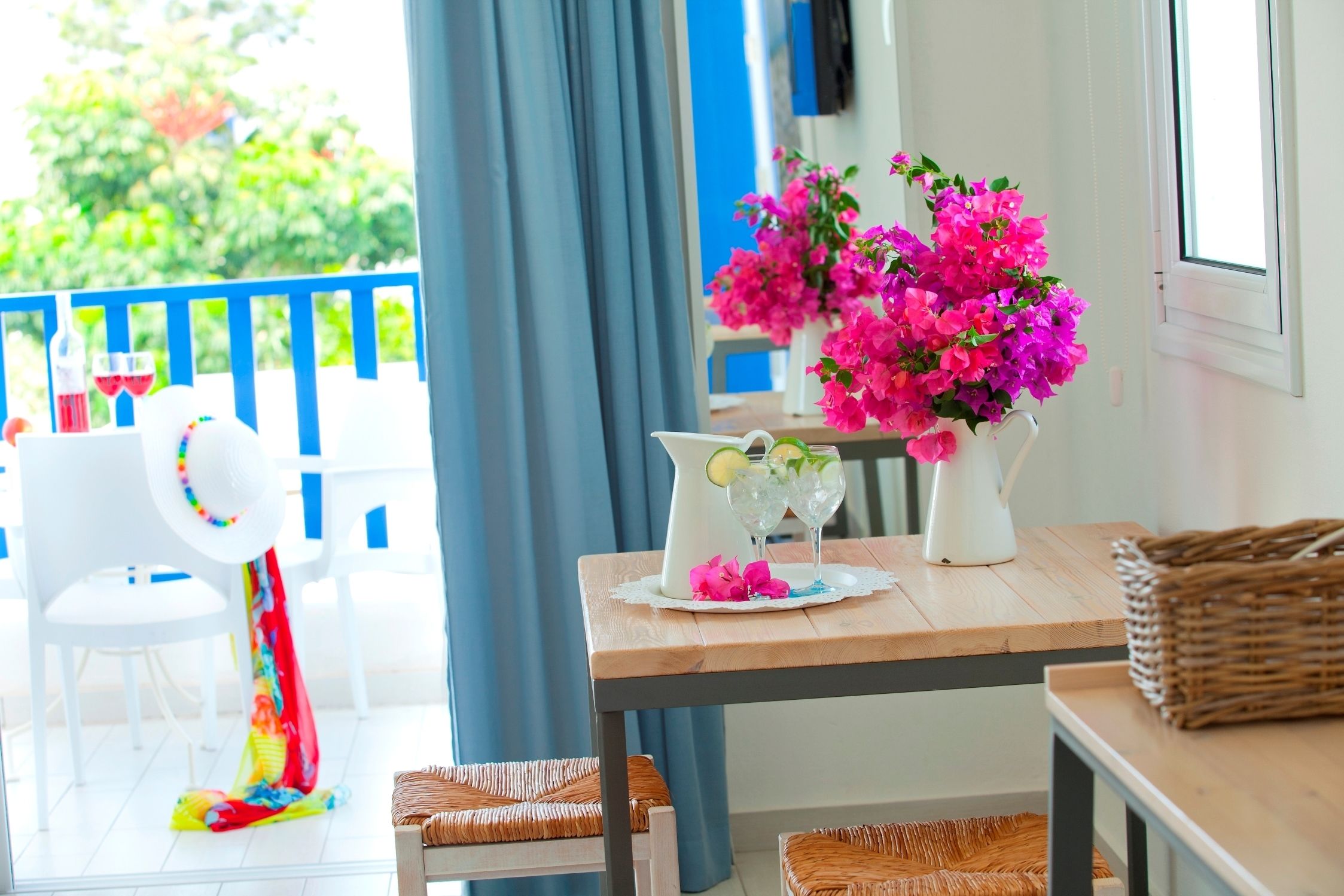 A bright room with a wooden table, wicker chairs, a white vase with pink flowers, and a wicker basket.