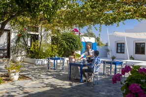 An outdoor seating area with tables and chairs, surrounded by potted plants and a white building with blue accents. - Aliathon Aegean (Geroskipou)
