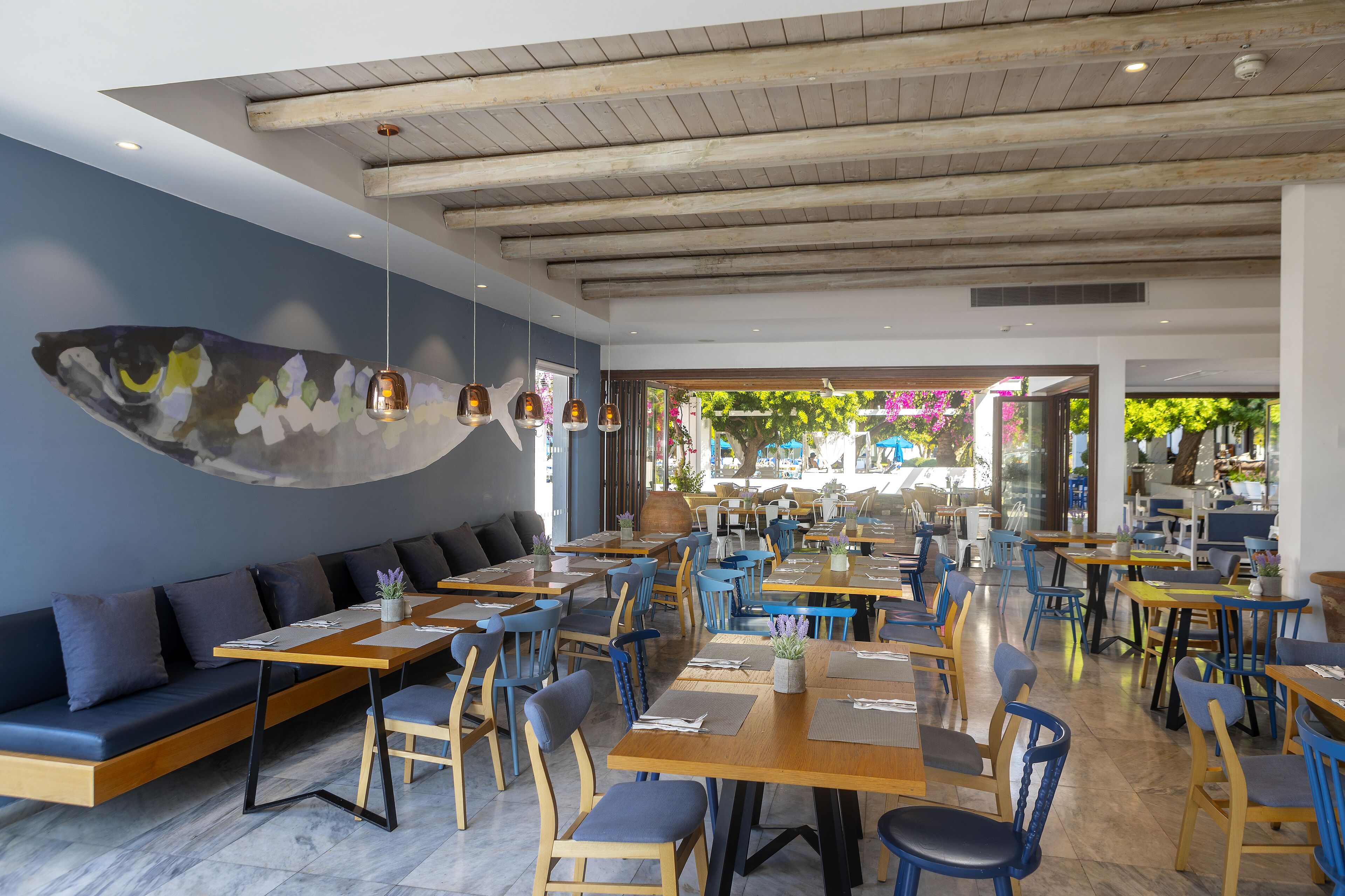 A modern restaurant with wooden tables and blue chairs, a large fish mural on the wall, and a view of the outdoors.