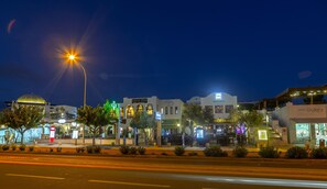 A street view at night with illuminated buildings, including a pharmacy, restaurant, and gallery. - Aliathon Aegean (Geroskipou)