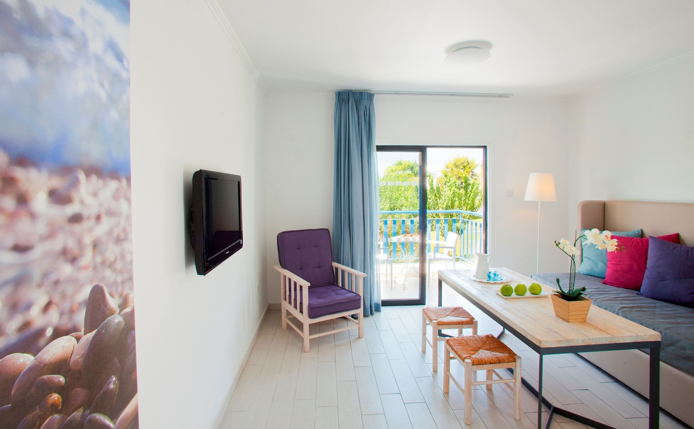 A modern room with a flat-screen TV mounted on the wall, a purple chair, a wooden table with a plant, and a sofa with colorful cushions.