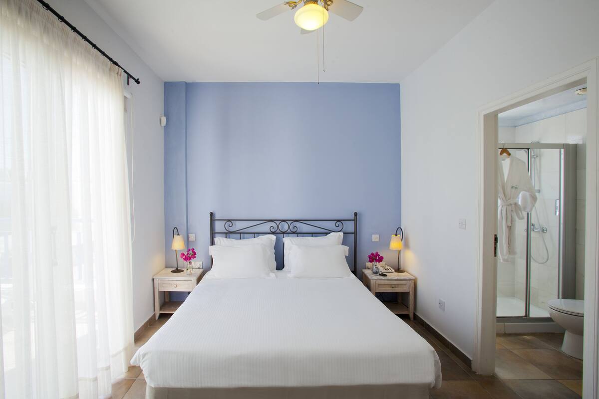 Habitación de hotel con cama, mesitas de noche, lámpara, espejo y zona de ducha.