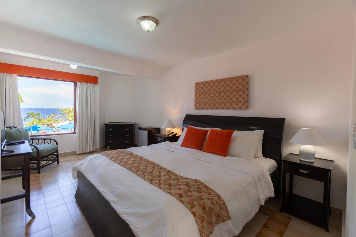 Ein Hotelzimmer mit einem großen Bett, einem Schreibtisch und einem Sessel. Es gibt ein Fenster mit Vorhängen, einen Nachttisch mit einer Lampe und ein Wandbild.