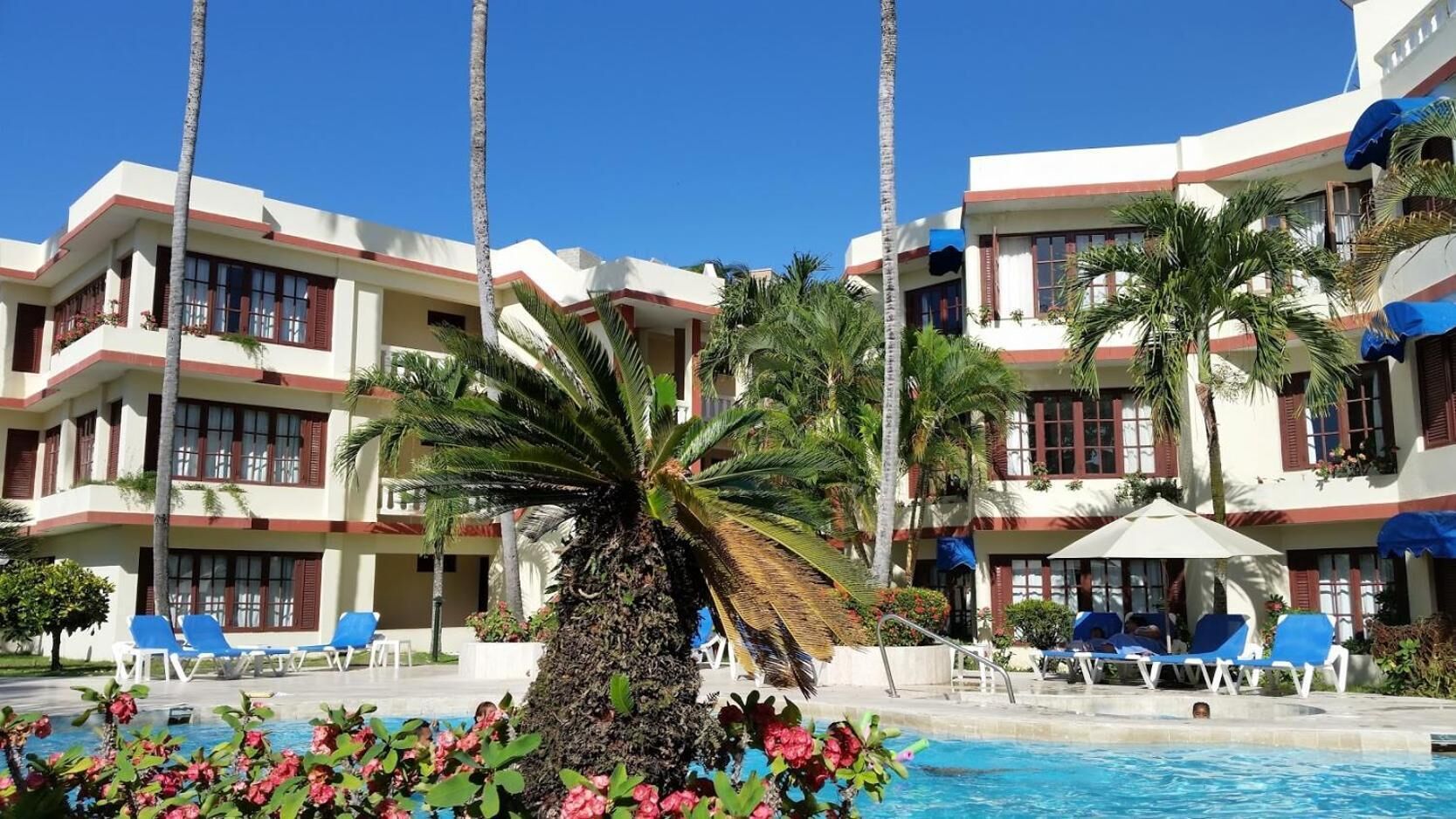Un hôtel doté d’une piscine, de palmiers et de chaises longues bleues.