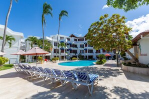 A resort with a swimming pool, lounge chairs, palm trees, and a restaurant.