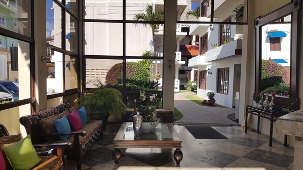 A spacious lobby with a checkered floor, a sofa with colorful cushions, a coffee table, and a view of the outdoor area with plants and a pathway.