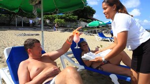 A beach scene with a person reclining on a blue lounge chair under a green umbrella, another person seated beside them, and a third person standing nearby. - Sosua by the Sea Boutique Beach Resort (Sosúa)