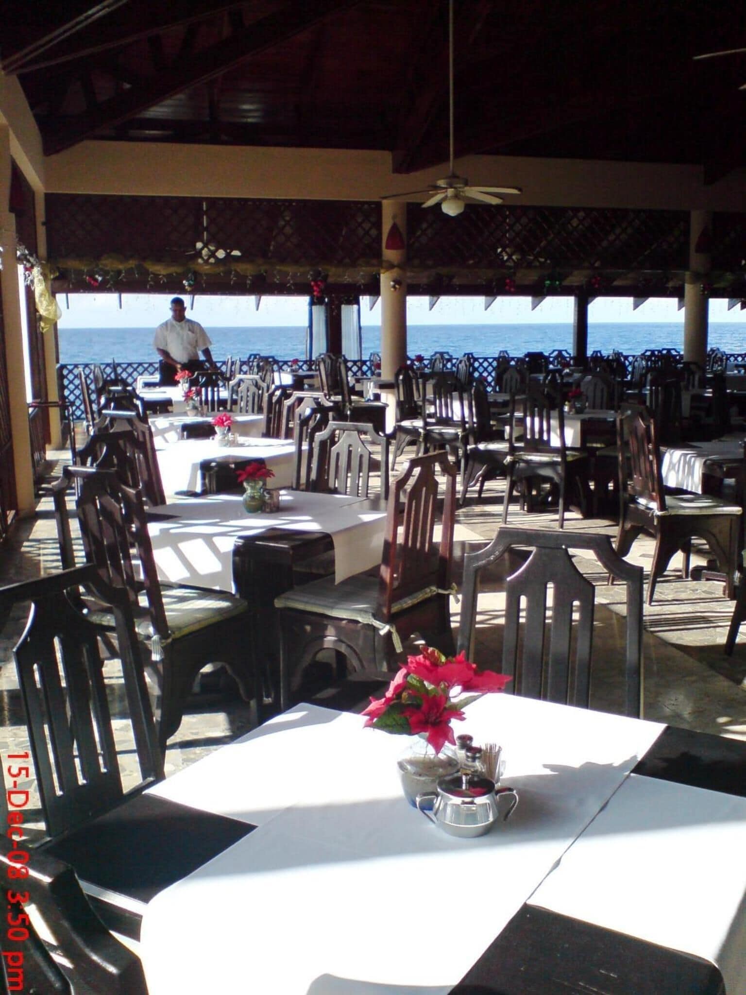 An outdoor dining area with tables and chairs, a chef in the background, and a view of the sea.