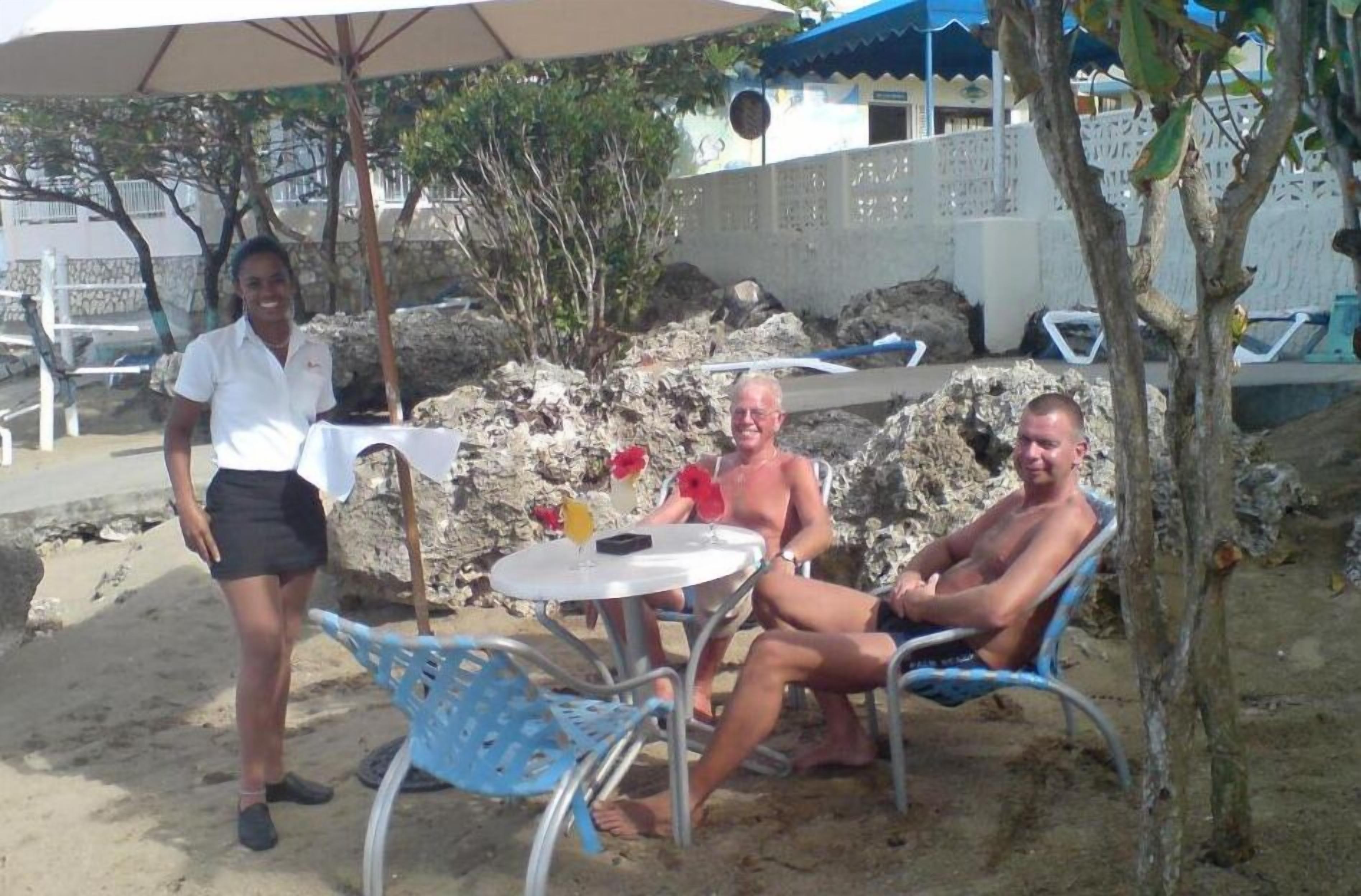 A beachside setting with a table and chairs under an umbrella, a man and woman seated, and a man lying on a lounge chair.