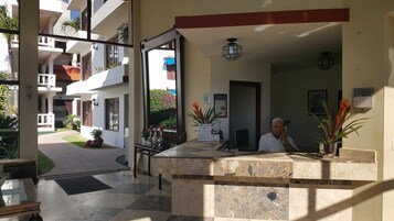 Zona de recepción de un hotel con recepcionista, mostrador de recepción y vistas al patio exterior.