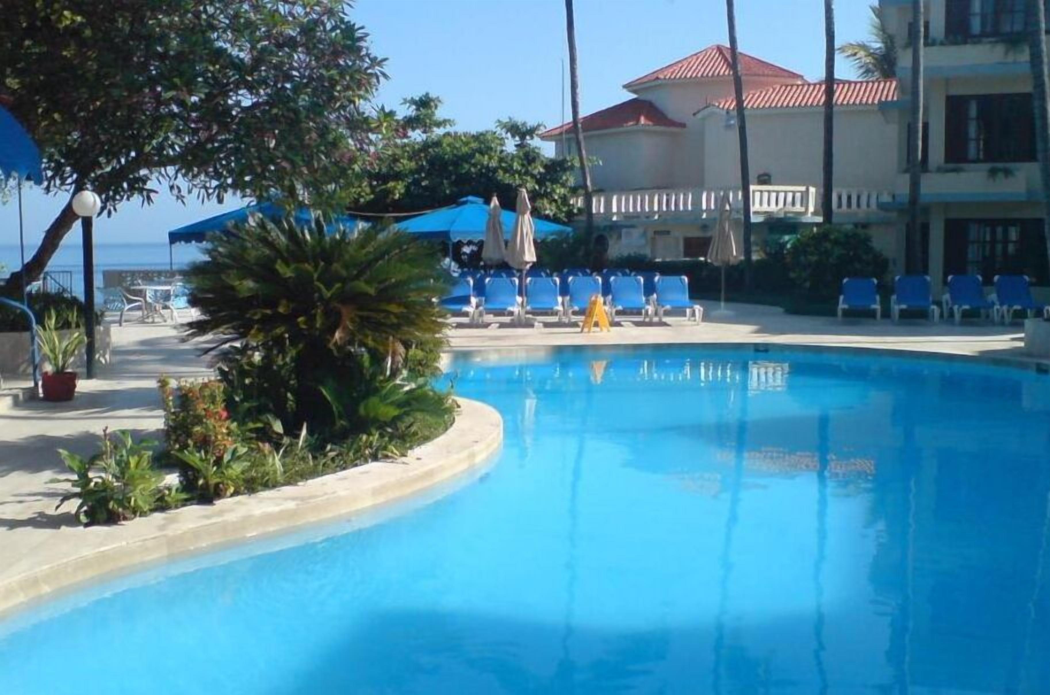 A swimming pool with blue chairs and umbrellas, surrounded by greenery and a building with a red roof.
