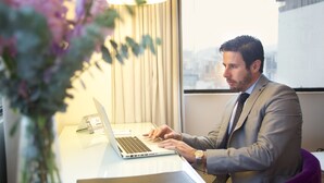 Un hombre con traje trabaja en una computadora portátil sobre un escritorio con una lámpara y un jarrón de flores.