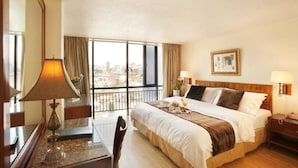 Habitación de hotel con una cama grande, un escritorio con lámpara, una silla, una mesita de noche y vistas a la ciudad a través de amplias ventanas.