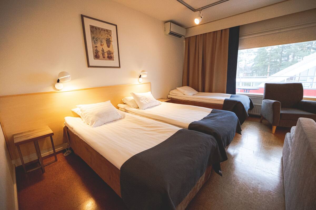 A hotel room with two single beds, a small table, a chair, and a window with curtains.