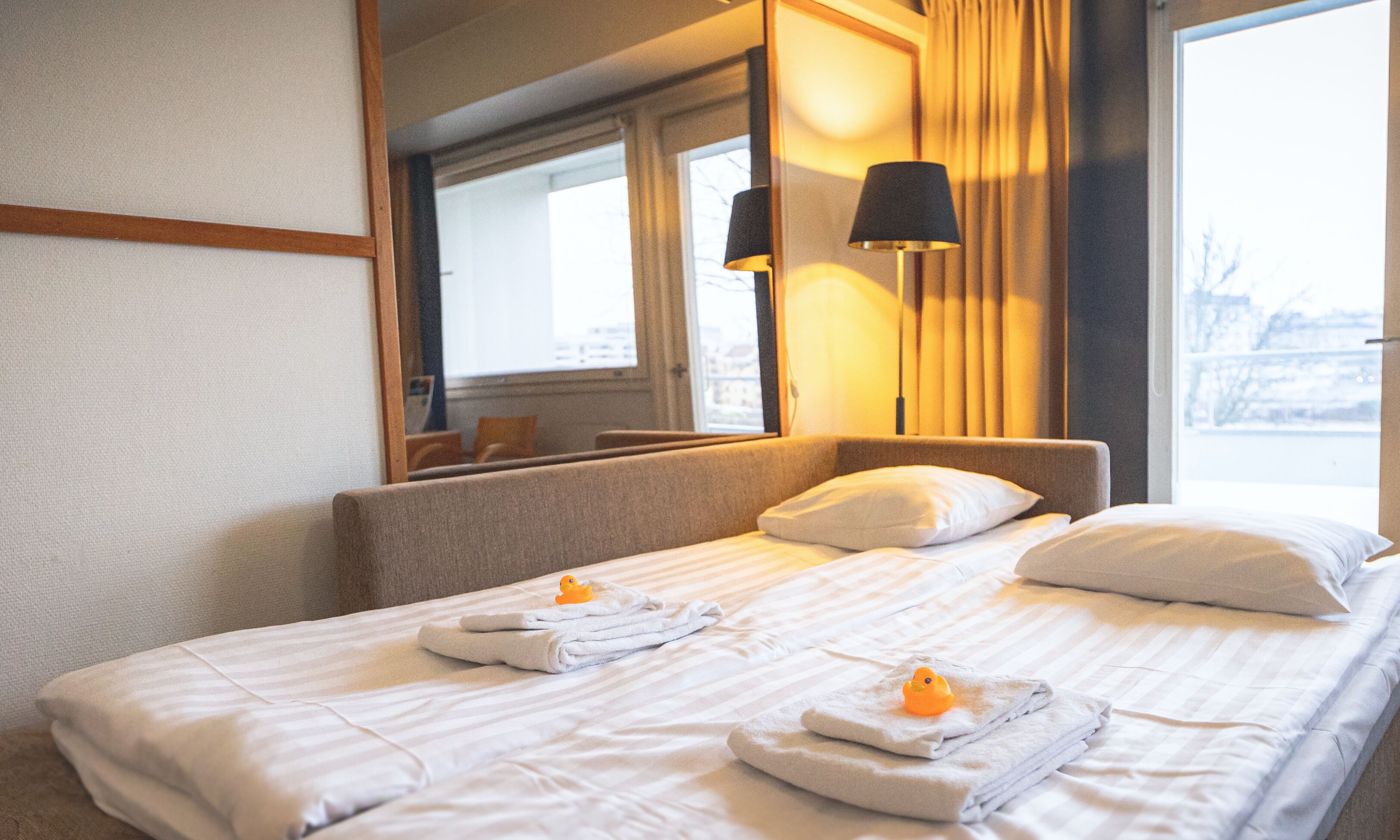 A hotel room with a neatly made bed, two pillows, a nightstand, a lamp, and a window with curtains.
