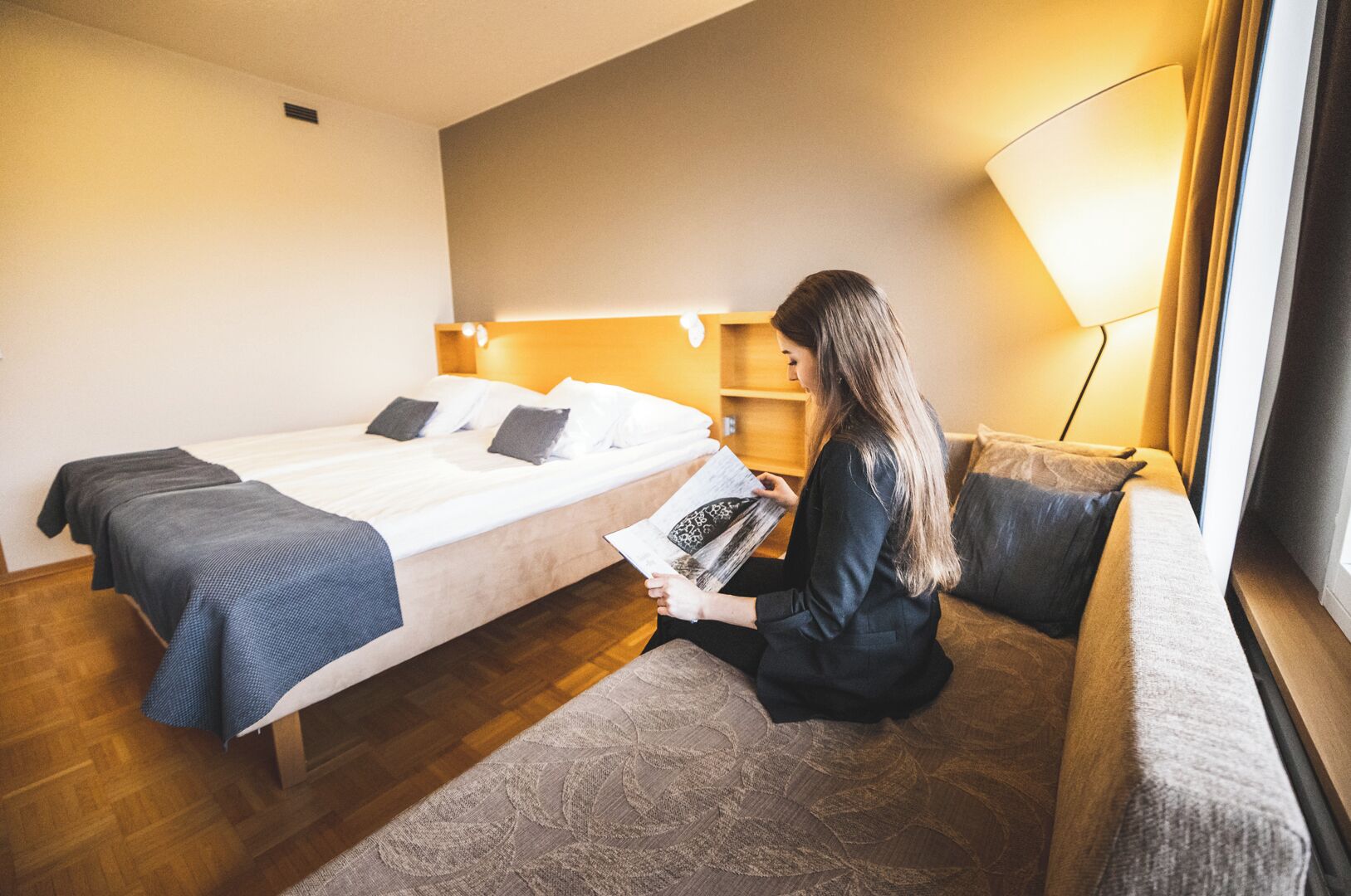 A hotel room with a bed, a sofa, a lamp, and a person reading a book.
