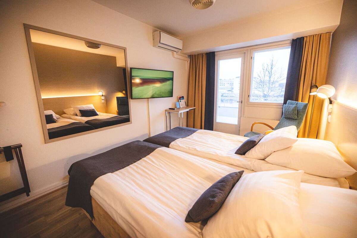 A hotel room with two beds, a TV, a desk, and a window with curtains.