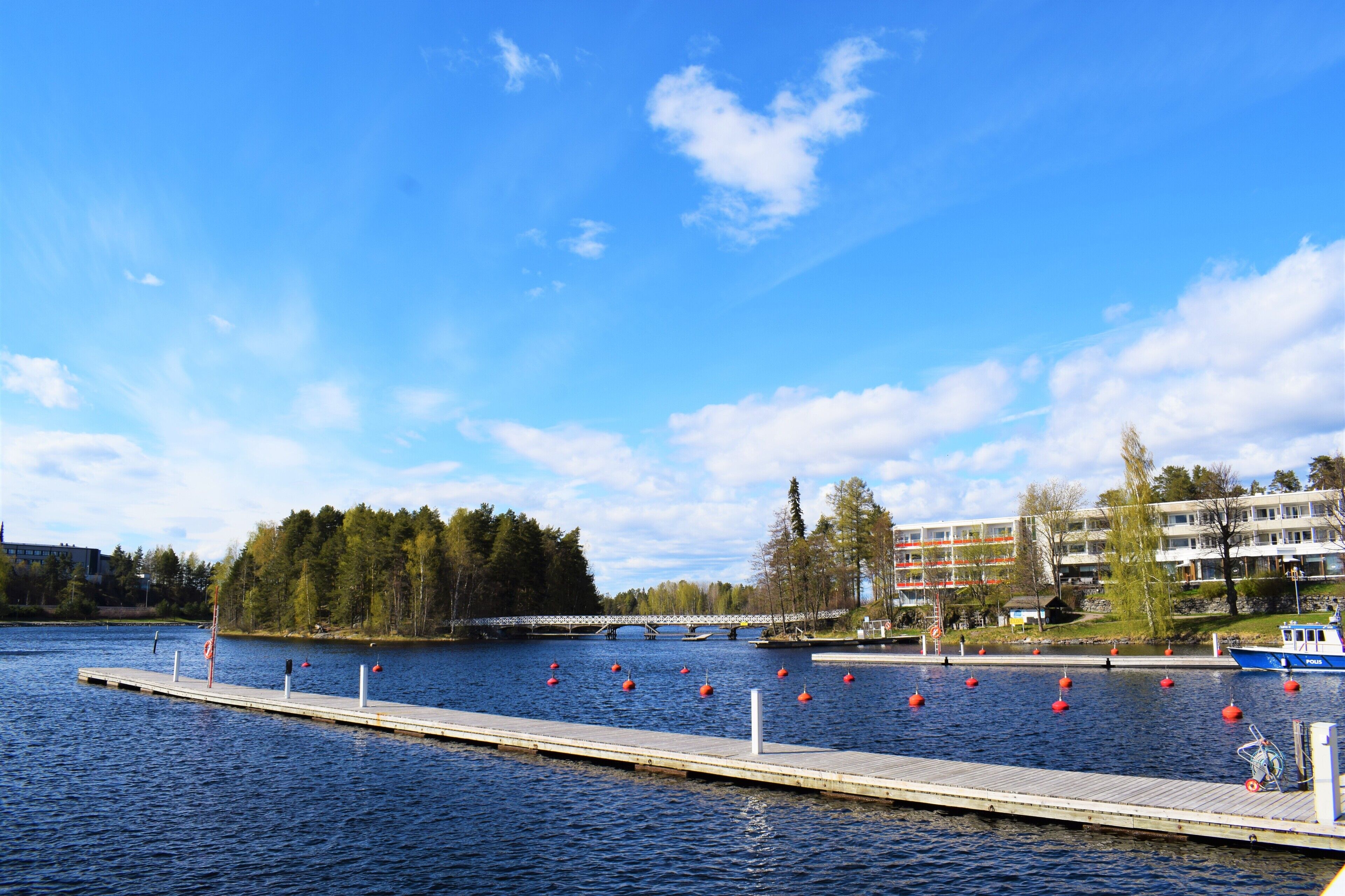 A calm lake with a wooden dock extending into the water, surrounded by trees and a building in the background.