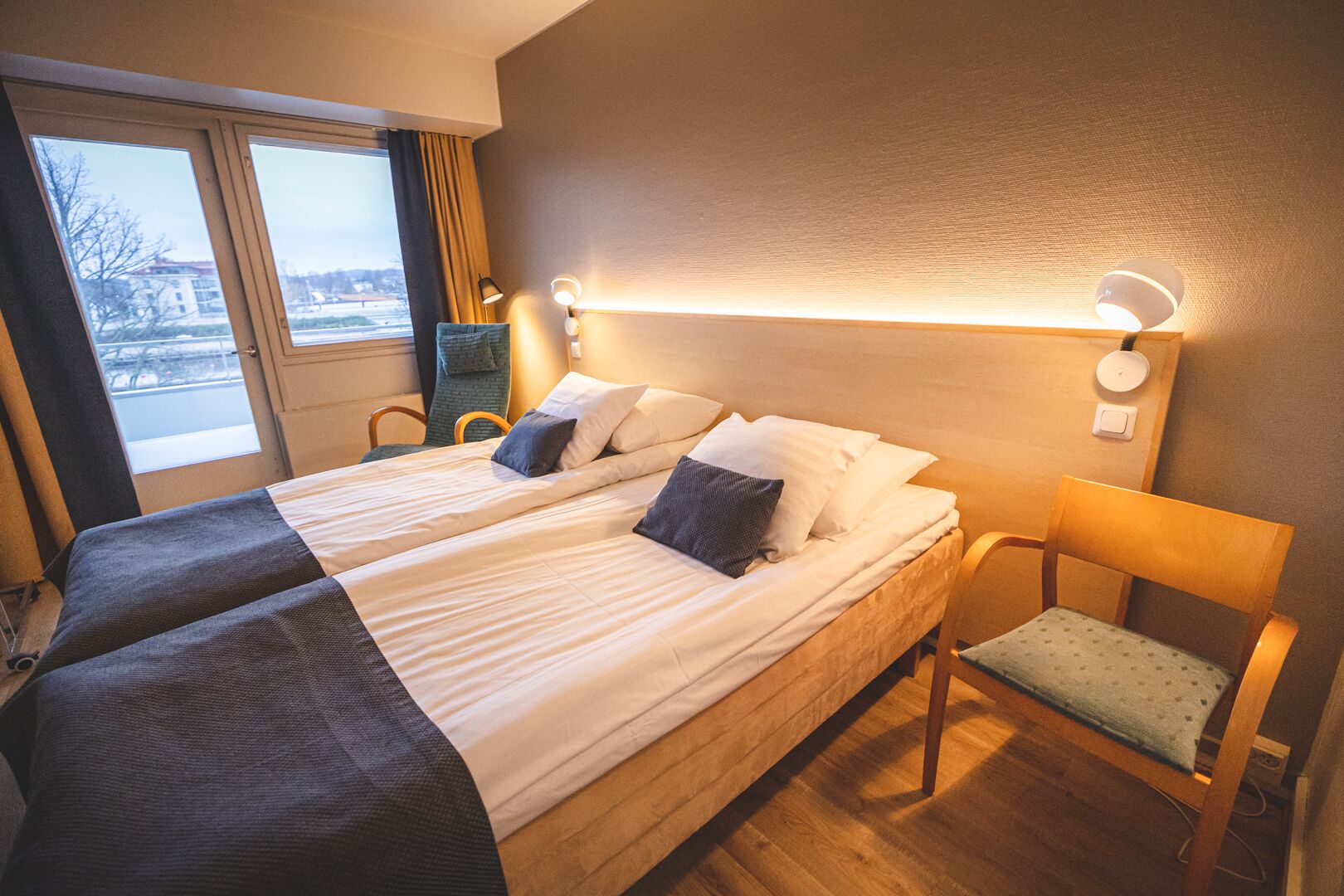 A double bed with two pillows, a bedside table with a lamp, and a chair with a cushion.