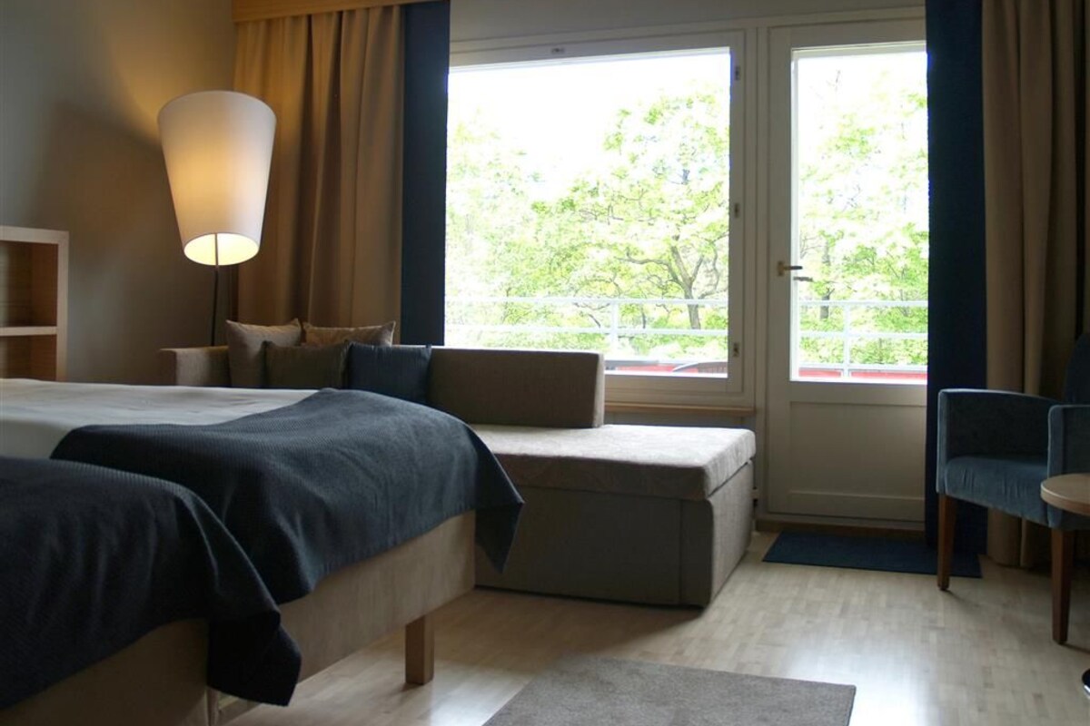 Hotellihuone, jossa on sänky, oleskelualue, työpöytä ja ikkuna, josta on näkymä vehreään maisemaan.