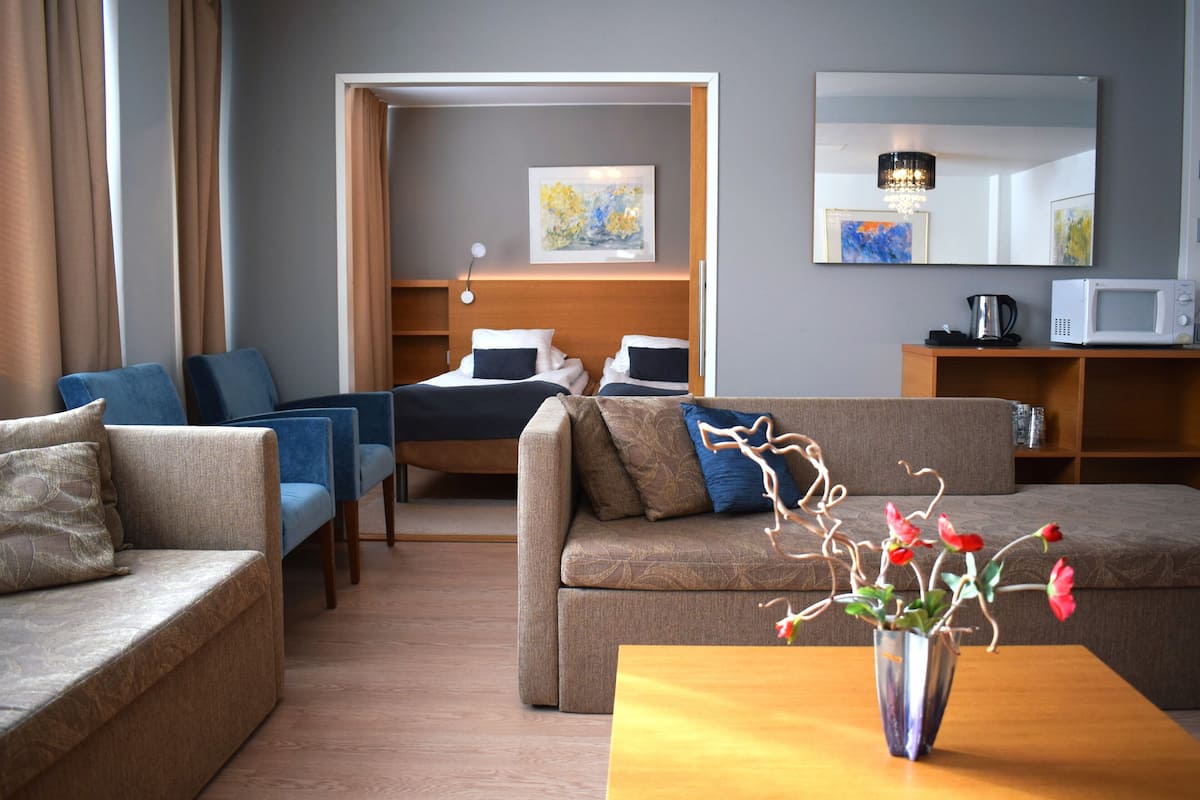 A hotel room with a sofa, a coffee table with a vase of flowers, a bed with a headboard, a bedside table, and a microwave on a shelf.