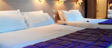 A hotel room with two beds, a purple quilt, and wall-mounted lamps.