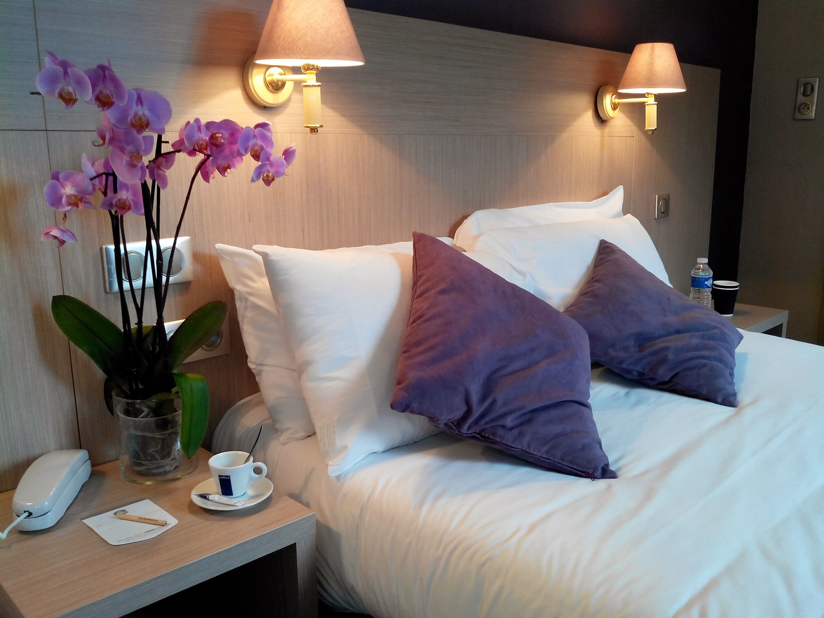 Habitación de hotel con una cama bien hecha, dos lámparas de pared, una mesita de noche con un teléfono, una taza, un bloc de notas y una orquídea en maceta.