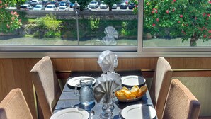 A table set for two with plates, cutlery, a pitcher, and a bowl of bread, overlooking a parking area and trees. - Hotel Christina Lourdes (Lourdes)