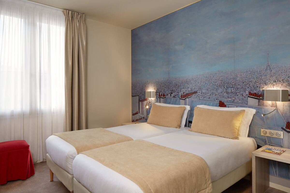 Une chambre d’hôtel avec deux lits, un pouf rouge, un tableau représentant un paysage urbain et une table de chevet avec un magazine.