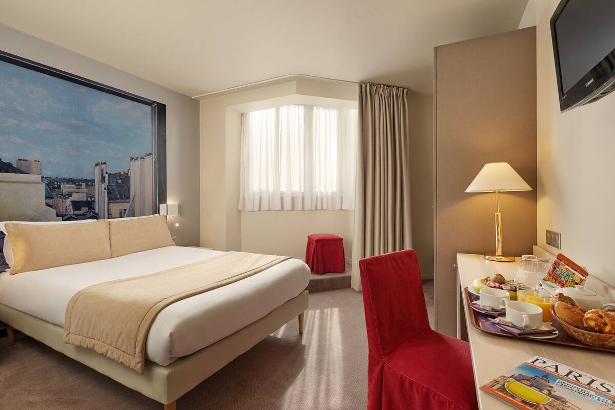 A hotel room with a bed, a desk with breakfast items, a chair, a window with curtains, and a painting on the wall.