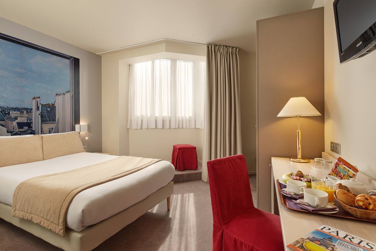 Une chambre d’hôtel avec un grand lit, un bureau avec des articles pour le petit-déjeuner, une chaise rouge, une fenêtre avec des rideaux et une vue sur la ville.