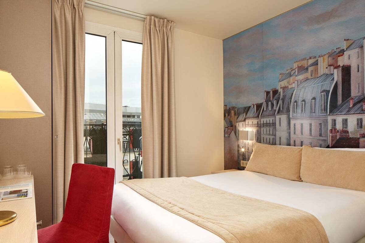 Une chambre d’hôtel avec un lit, un fauteuil rouge, une lampe, un tableau représentant des bâtiments et une vue sur l’extérieur.