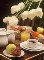 A breakfast table with croissants, a fruit basket, a pitcher, a glass of orange juice, and a cup of coffee.