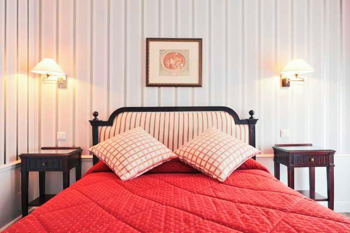Un lit bien fait, recouvert d’une couette rouge, avec deux oreillers assortis et deux tables de chevet dotées de lampes.